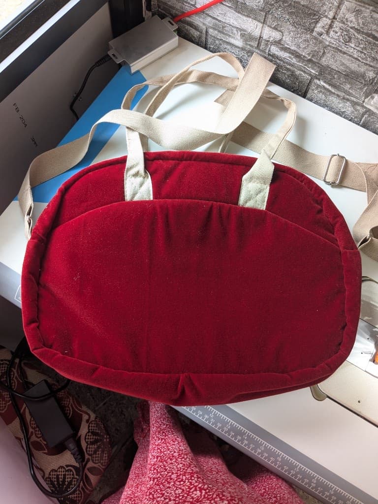 Back view of the padded burgundy velvet diaper bag on the workshop table showing the pillowed panel and cream webbing straps