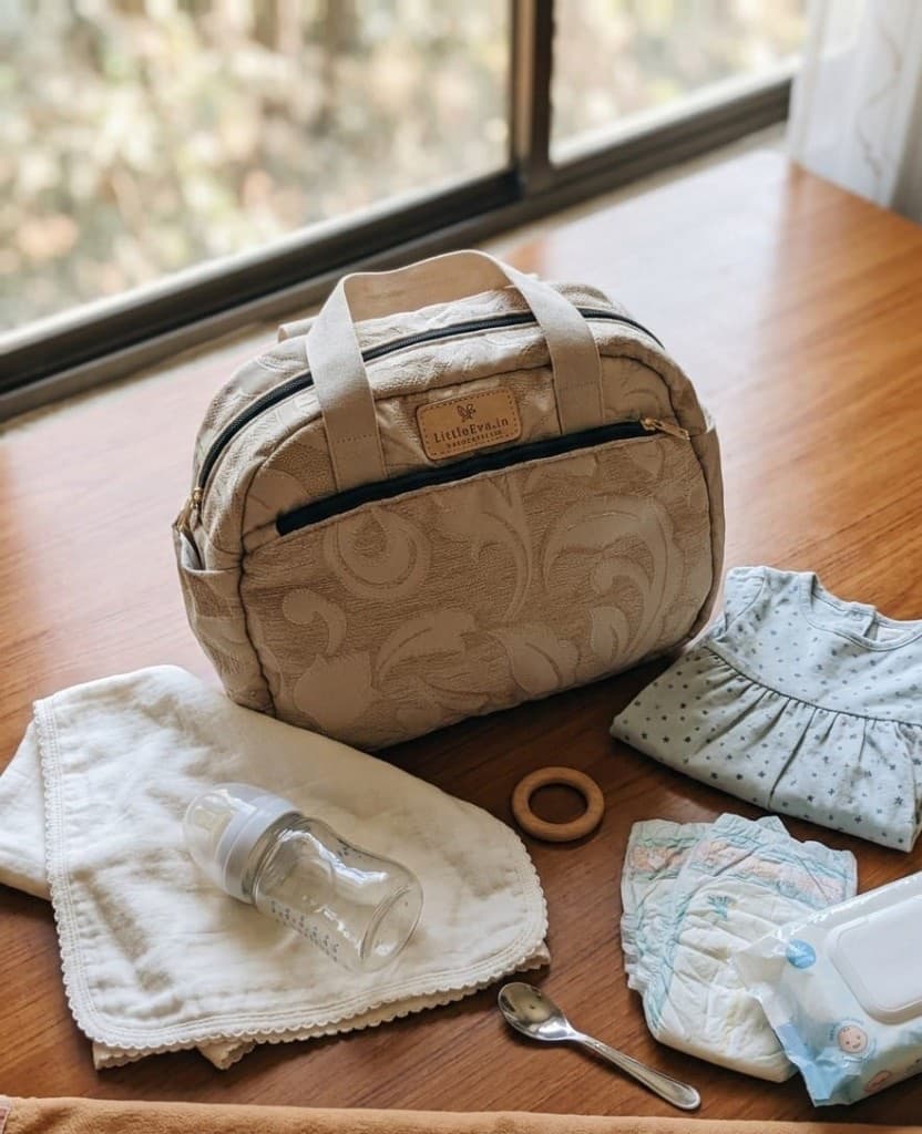 Cream jacquard diaper bag styled on a wooden table with baby bottle, diapers, wipes, and a muslin cloth