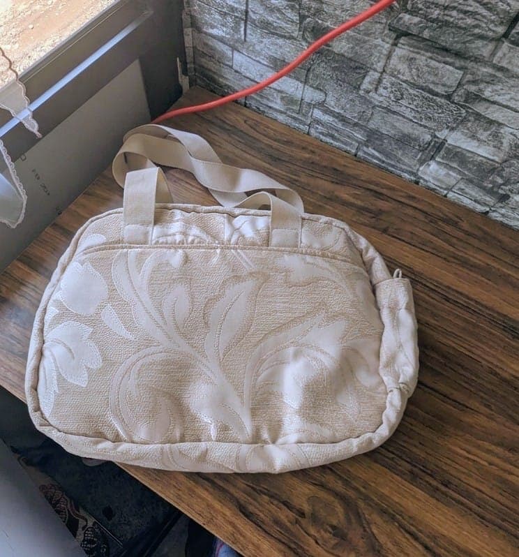 Cream jacquard diaper bag on a wooden worktable showing piping, front pocket, and elastic side pocket